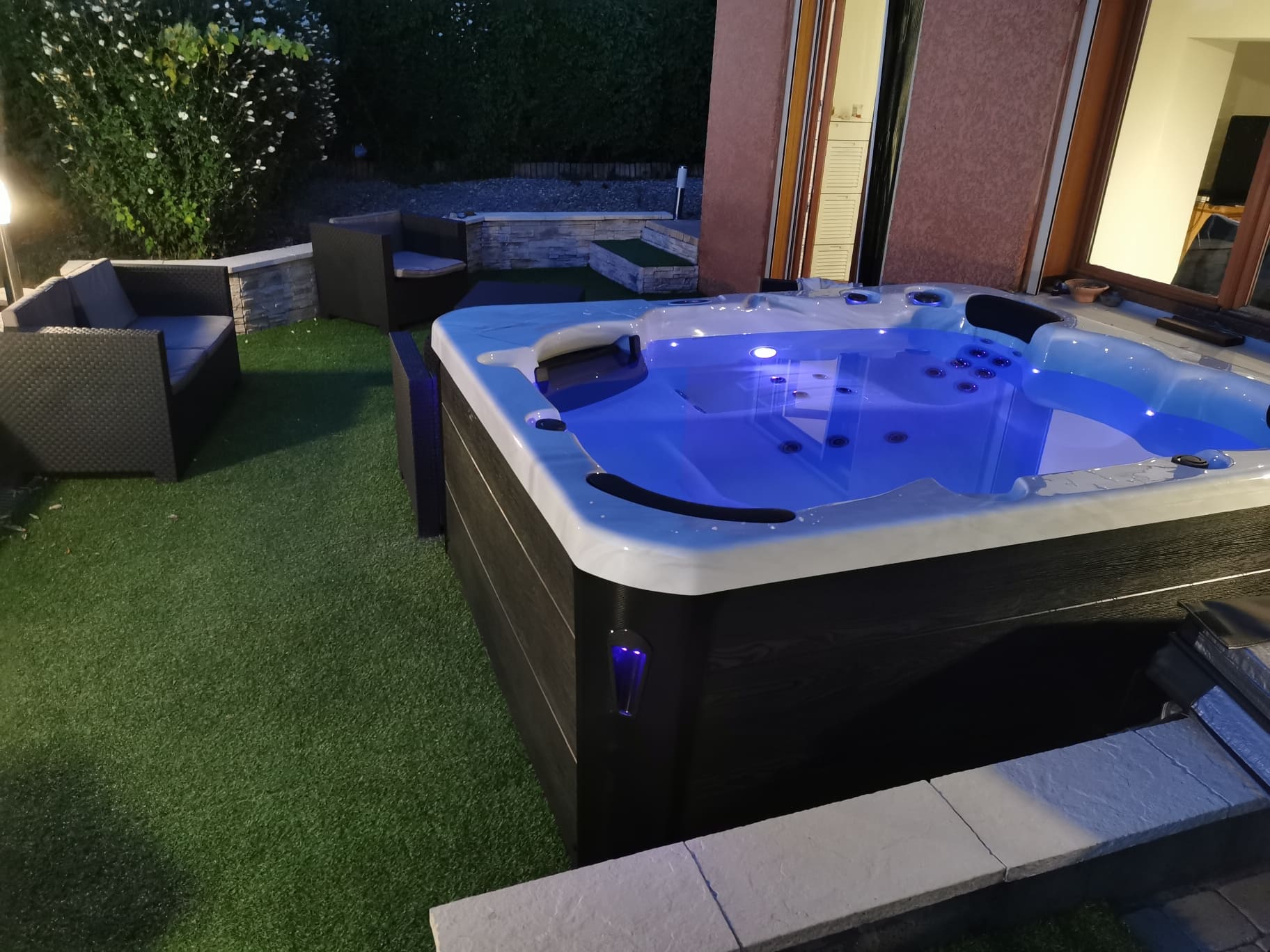 Spa de massage extérieur éclairé par des leds bleues, installé sur gazon synthétique avec salon de jardin cosy en arrière-plan, de nuit.